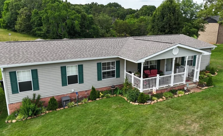 A one-story gray house with green shutters and a white porch with red chairs, surrounded by a green lawn.